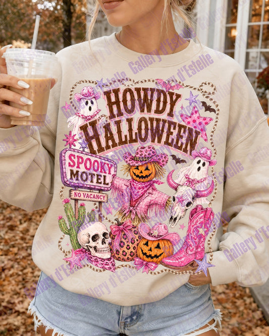 A woman wearing a cream-colored sweatshirt with a colorful 'Howdy Halloween Spooky Motel' graphic print, holding an iced coffee outdoors in front of a porch with pumpkins.