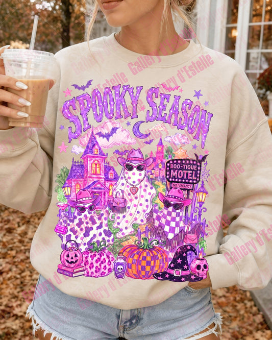 A woman wearing a beige crewneck sweatshirt with a colorful 'Spooky Season' graphic print featuring three country ghosts, a haunted house, pumpkins, and bats. She is holding an iced coffee outdoors in a fall setting.