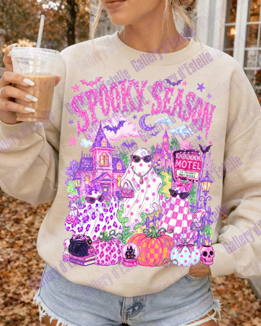 A woman wearing a beige crewneck sweatshirt with a colorful 'Spooky Season' graphic featuring three glam ghosts, haunted house, pumpkins, and purple leopard print details, holding an iced coffee outdoors.