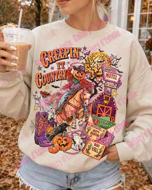 A woman wearing a cream-colored crewneck sweatshirt with a colorful 'Creepin' in the Country' Halloween graphic, holding an iced coffee outdoors.