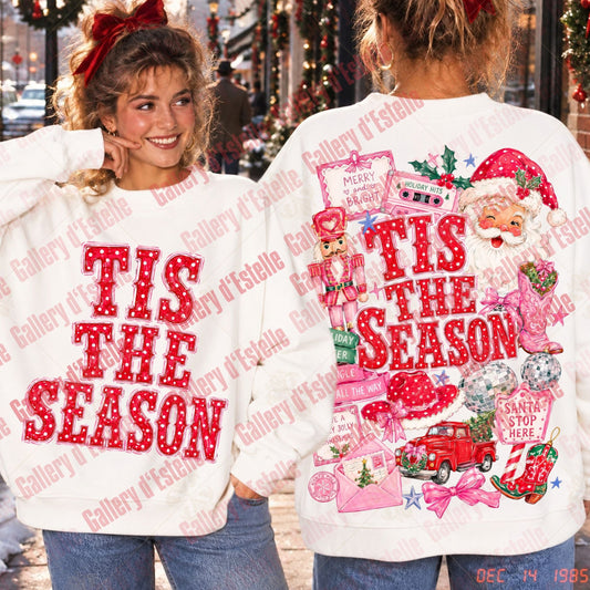Two women modeling white Christmas crewneck sweatshirts in a festive outdoor setting. The front view shows 'TIS THE SEASON' in red glitter-style letters, while the back view features a large, colorful holiday collage with Santa, a nutcracker, a red truck, and 'TIS THE SEASON' text.