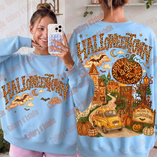 A light blue crewneck sweatshirt shown on a model from both the front and back. The front features a leopard-print 'HALLOWEENTOWN' text graphic, while the back displays a detailed Halloween-themed illustration with a haunted house, yellow car and pumpkins