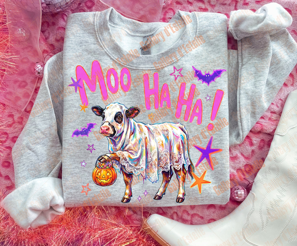 A light heather gray sweatshirt laid flat on a pink textured surface, featuring a colorful graphic of a cow in a Halloween dress with the text 'Moo Ha Ha!' and stars, bats, and pumpkins. White cowboy boots and sheer ribbon are styled around it.