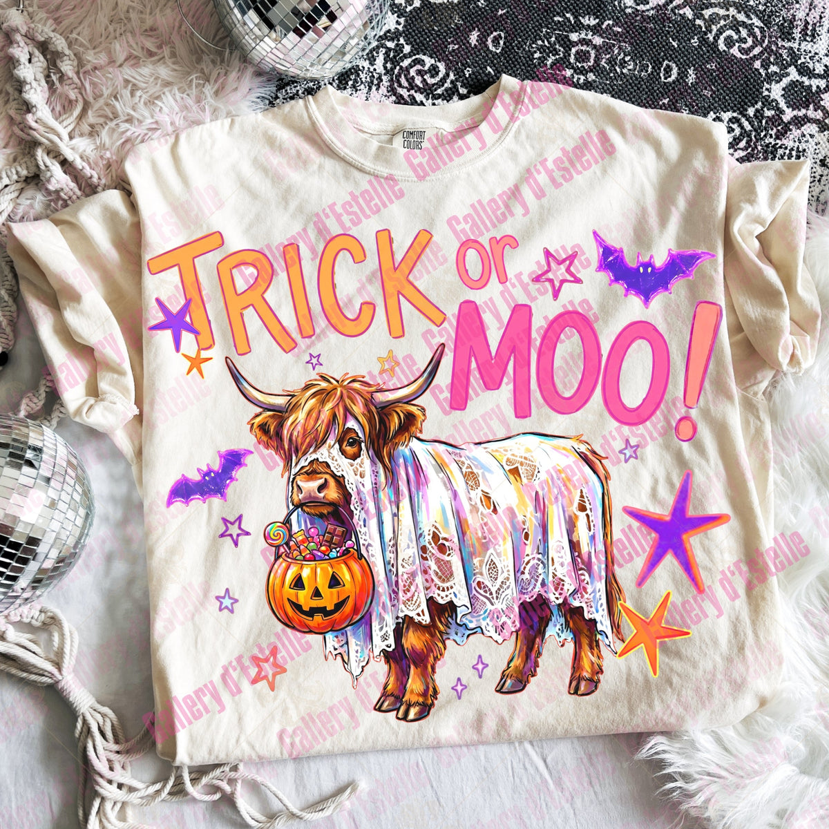 A cream-colored t-shirt featuring a colorful graphic of a Scottish highland cow wearing a lace dress and holding a jack-o'-lantern, surrounded by purple bats, orange stars, and the text 'TRICK or MOO!'. The shirt is displayed in a flat-lay arrangement on a white faux fur surface with a black lace cloth, a disco ball, and a silver beaded bag as props.