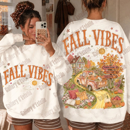 Two women modeling matching white sweatshirts with 'FALL VIBES' text and autumn graphics. One shows the front with orange lettering, and the other shows the back with a detailed fall harvest scene including a pickup truck, house, pumpkins, and signs.