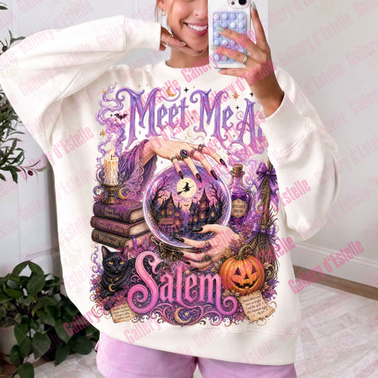 A woman takes a mirror selfie wearing a cream-colored sweatshirt with a large purple and pink Halloween graphic that reads 'Meet Me A Salem' with a crystal ball, witch hat, and pumpkin.