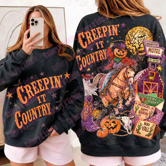 A woman modeling a black graphic sweatshirt with two views: a mirror selfie showing the front with orange 'CREEPIN' IT COUNTRY' text, and a back view showing a large Halloween-themed country western graphic with a horse, barn, pumpkins, and signs.