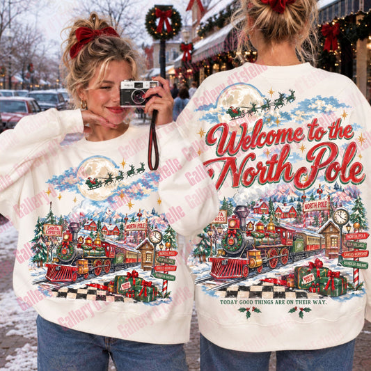Two women modeling a white Christmas sweater, showing both the front and back designs. The front features a small holiday village scene with Santa in a sleigh, while the back displays a large red 'Welcome to the North Pole' text above a detailed festive village and train illustration.