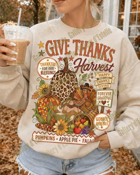 A woman wearing a cream-colored crewneck sweatshirt with a colorful 'Give Thanks Harvest' Thanksgiving graphic, holding an iced coffee in an outdoor autumn setting.