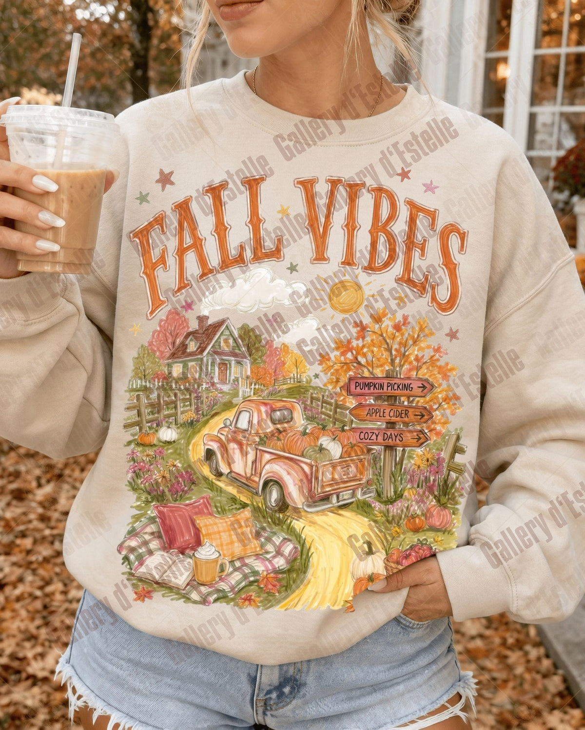 A woman wearing a cream-colored crewneck sweatshirt with a colorful 'Fall Vibes' graphic print featuring an orange pickup truck, autumn house, and pumpkins. She is holding an iced coffee in a real outdoor setting with fallen leaves on the ground.