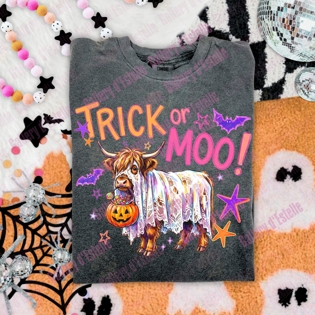 A dark gray graphic t-shirt featuring a cartoon Highland cow draped in a lace nightgown, holding a jack-o'-lantern, with the text 'TRICK or MOO!' in orange and pink. The shirt is displayed in a flat-lay arrangement with Halloween-themed props including a spiderweb, beads, a disco ball, and a ghost blanket.