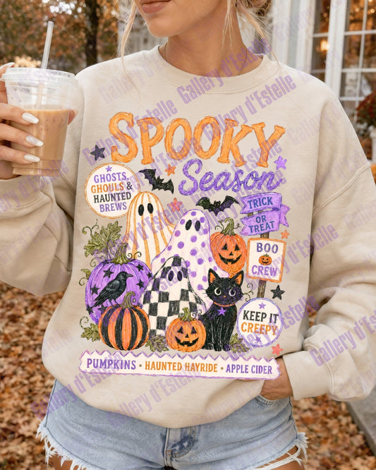 A woman wearing a cream-colored crewneck sweatshirt with a colorful 'Spooky Season' graphic print, holding an iced coffee in an outdoor autumn setting.