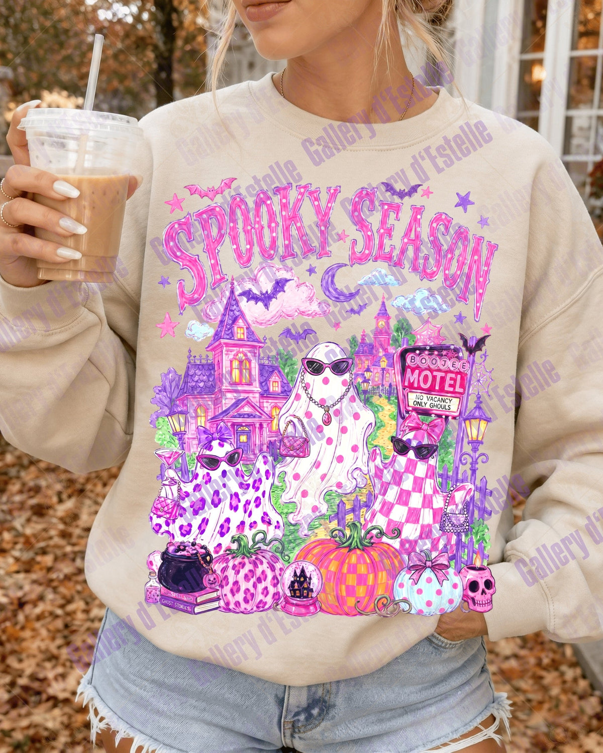A woman wearing a beige crewneck sweatshirt with a colorful 'Spooky Season' graphic featuring three glam ghosts, haunted house, pumpkins, and purple leopard print details, holding an iced coffee outdoors.
