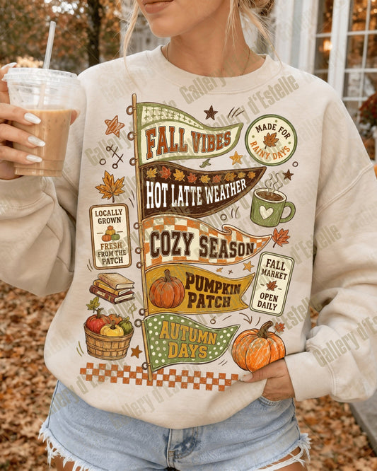 A woman wearing a cream-colored crewneck sweatshirt with a colorful autumn-themed graphic print that reads 'Fall Vibes' and 'Cozy Season' with pumpkins, leaves, and coffee cups, holding an iced coffee in an outdoor fall setting.