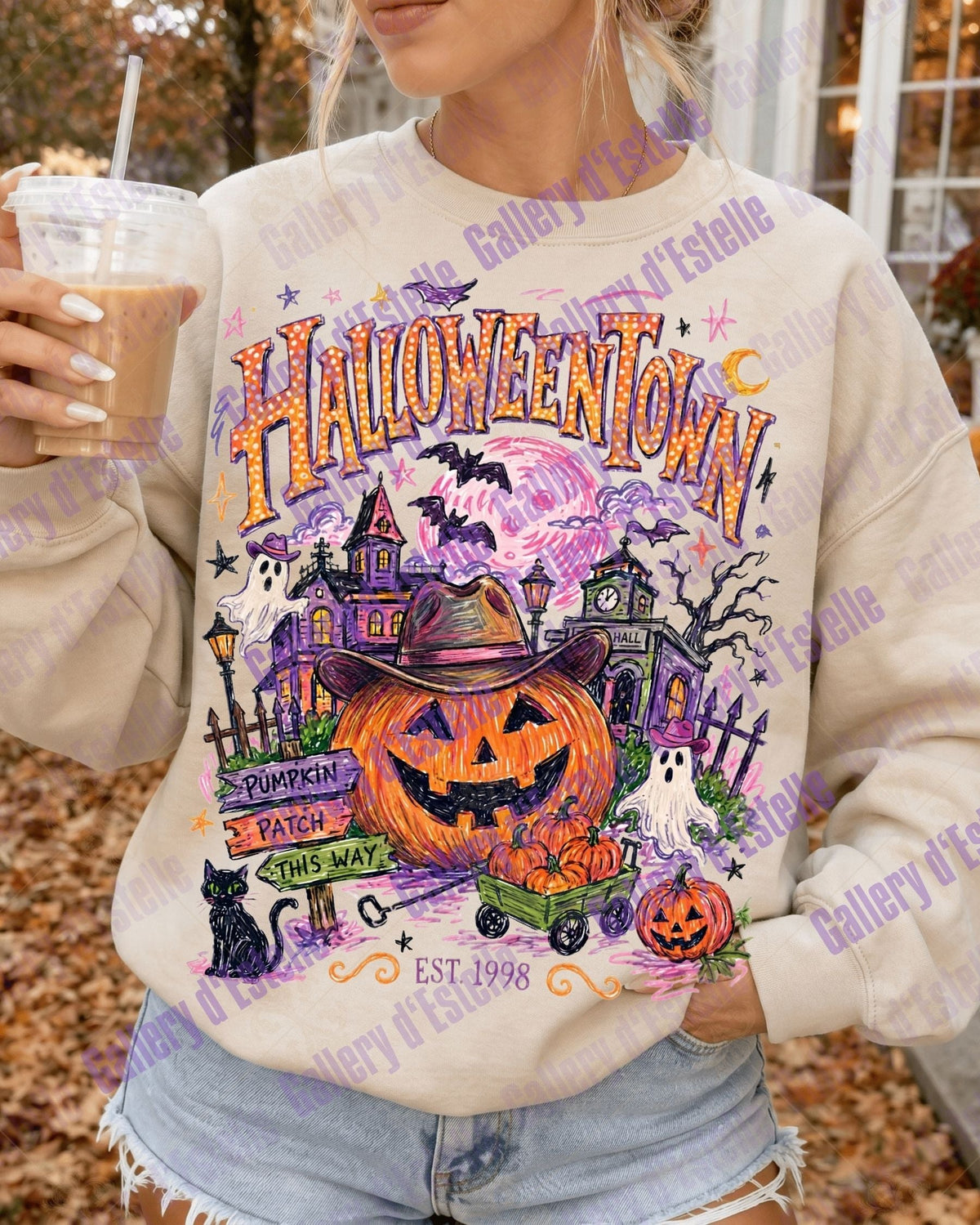 A woman wearing a cream-colored crewneck sweatshirt with a colorful 'Halloween Town' graphic print featuring a jack-o'-lantern, haunted house, and pumpkins, holding an iced coffee outdoors.