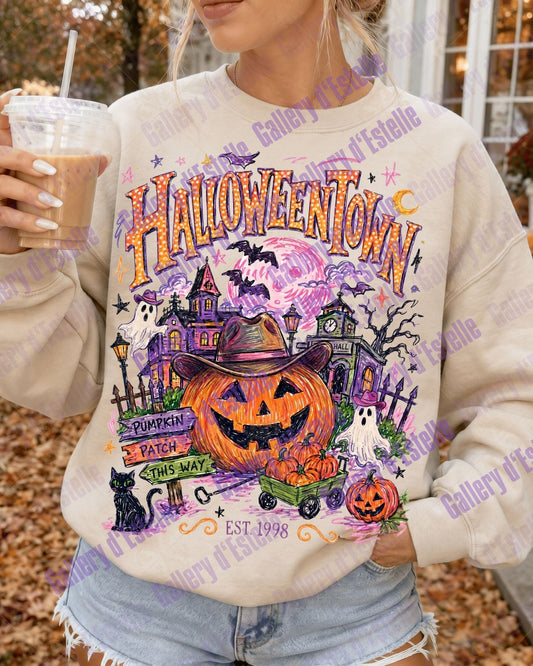 A woman wearing a cream-colored crewneck sweatshirt with a colorful 'Halloween Town' graphic print featuring a jack-o'-lantern, haunted house, and pumpkins, holding an iced coffee outdoors.