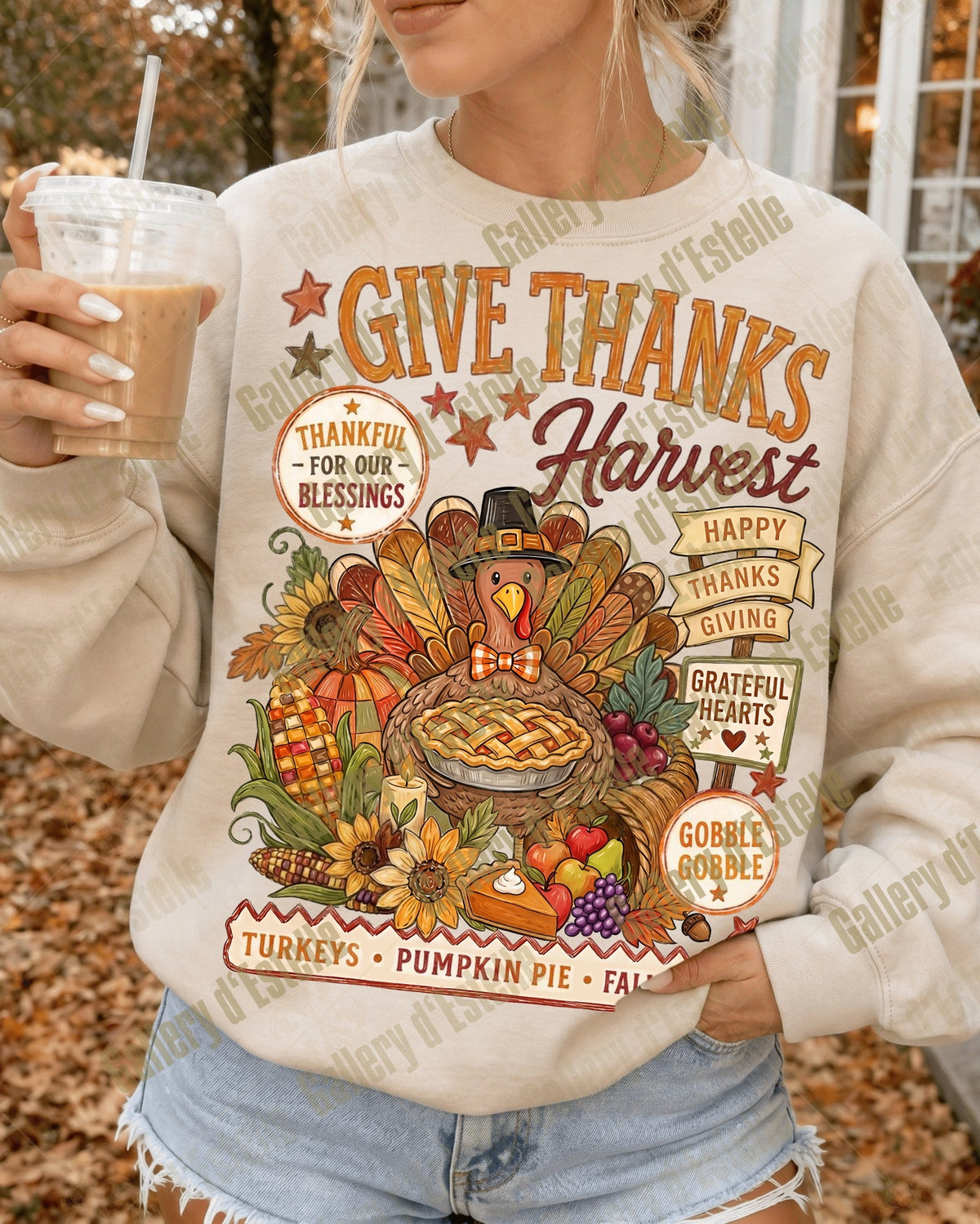 A woman wearing a cream-colored crewneck sweatshirt with a colorful autumn Thanksgiving graphic, holding an iced coffee in an outdoor fall setting.