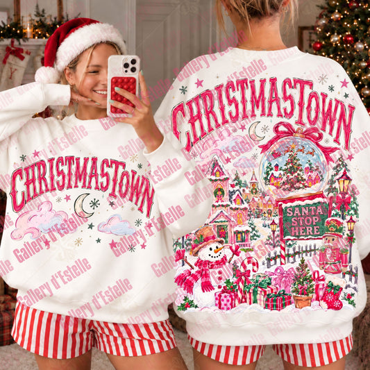 A person shown from the back wearing a white Christmas-themed sweatshirt with large pink 'CHRISTMASTOWN' text and a colorful snow globe village graphic, paired with red-and-white striped shorts. A mirror selfie view on the left shows the front of the same sweatshirt with matching pink text and holiday graphics.