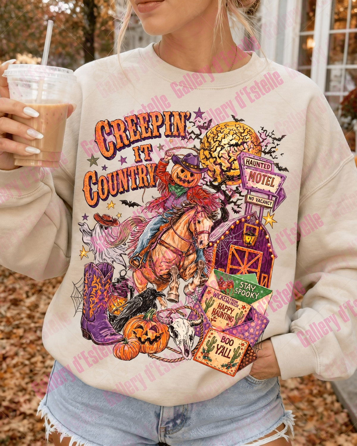 A woman wearing a cream-colored crewneck sweatshirt with a colorful 'Creepin' in the Country' Halloween graphic, holding an iced coffee outdoors.