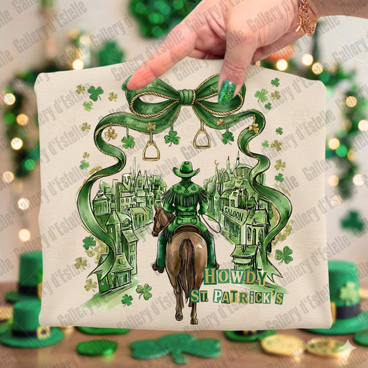 Beige folded T-shirt with a cowboy and saloon design, featuring shamrock patterns and 'Howdy St. Patrick's' text, held by a hand.