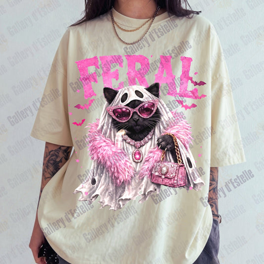 A person wearing an oversized cream-colored graphic t-shirt featuring a stylized black cat dressed in pink sunglasses, a white veil, and a pink fluffy boa under the word 'FERAL' in pink. 