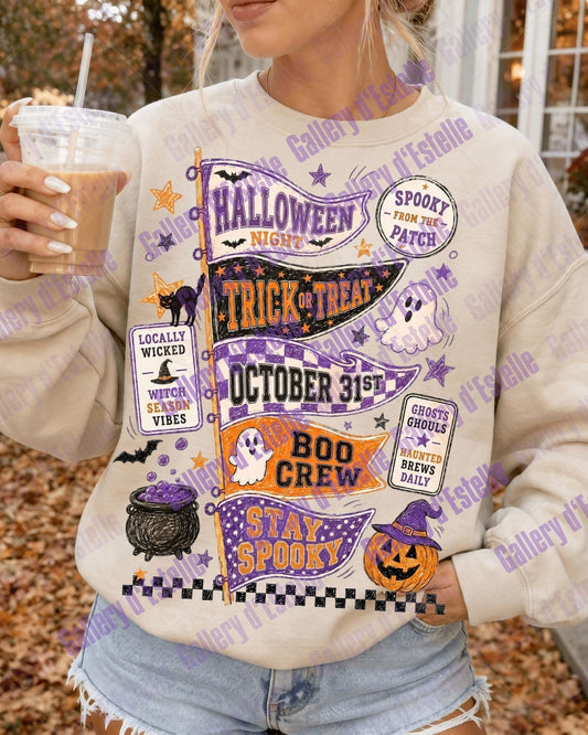 A woman wearing a cream-colored crewneck sweatshirt with a colorful Halloween-themed graphic print featuring purple, orange, and black text and illustrations, holding an iced coffee in an outdoor autumn setting.