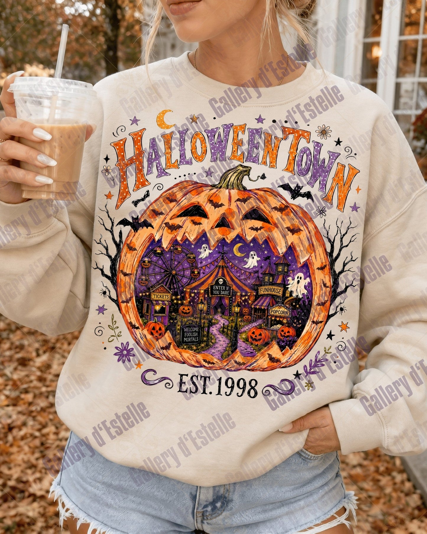 A woman wearing a cream-colored sweatshirt with a colorful 'Halloween Town' pumpkin graphic, holding an iced coffee outdoors.