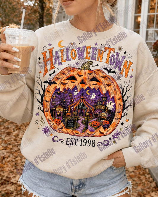 A woman wearing a cream-colored sweatshirt with a colorful 'Halloween Town' pumpkin graphic, holding an iced coffee outdoors.