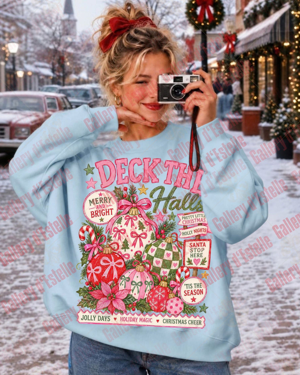 A woman takes a mirror selfie wearing a light blue crewneck sweatshirt with a colorful Christmas graphic that reads 'DECK THE HALLS' and features holiday ornaments, candy canes, and snow. The background shows a snowy street with decorated storefronts.