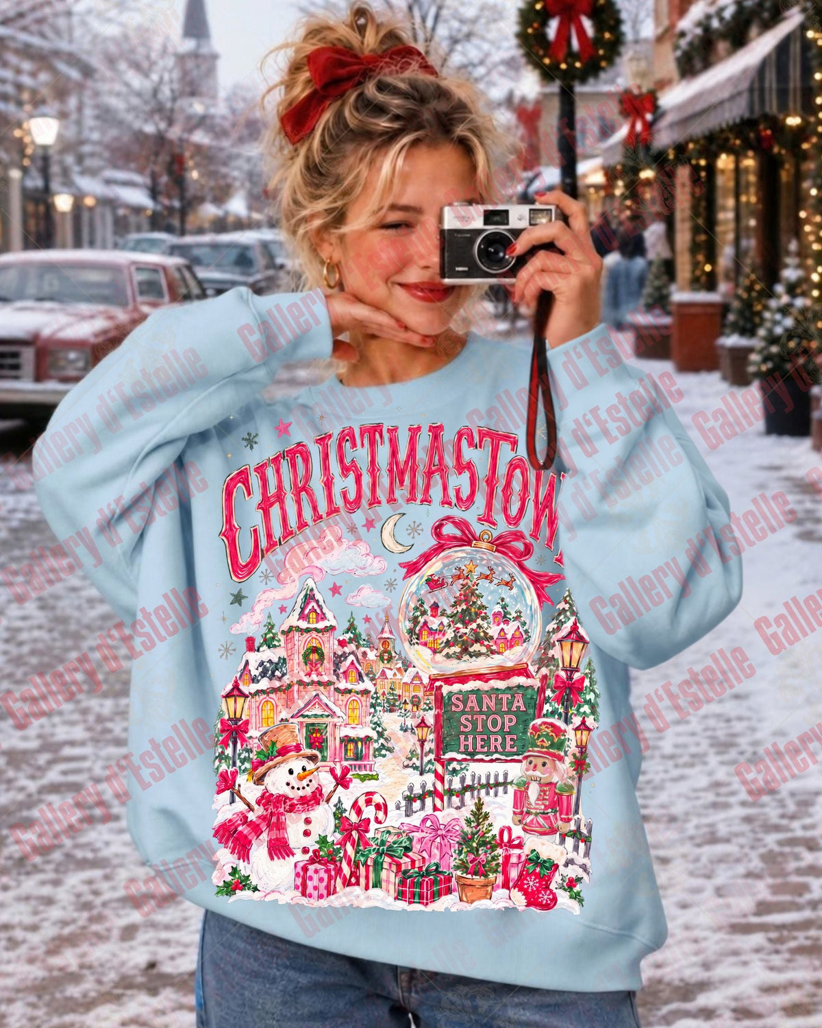 A woman wearing a light blue Christmas-themed sweatshirt with a festive village graphic and 'CHRISTMAS TOWN' text, taking a mirror selfie in a snowy outdoor storefront setting.