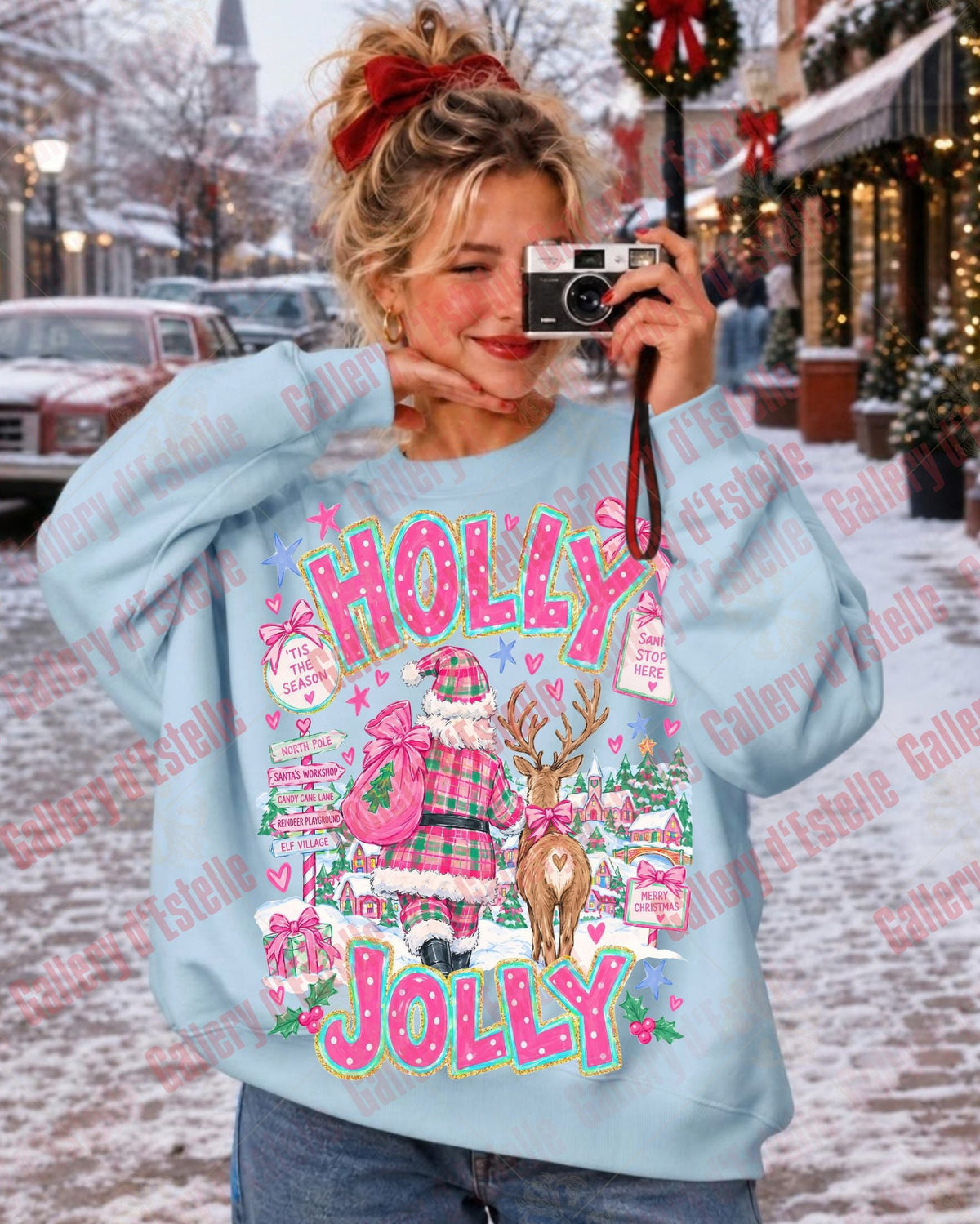 A woman takes a mirror selfie wearing a light blue crewneck sweatshirt with a colorful Christmas graphic that reads 'HOLLY JOLLY' and features Santa, a reindeer, and holiday motifs. The background shows a snowy street scene with storefronts and Christmas decorations.