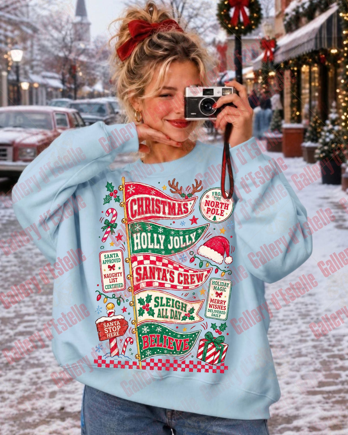 A woman takes a mirror selfie in a snowy outdoor street setting, wearing a light blue crewneck sweatshirt with a large, colorful Christmas-themed graphic print featuring text like 'Holly Jolly', 'Santa's Crew', and 'Sleigh to Go'.