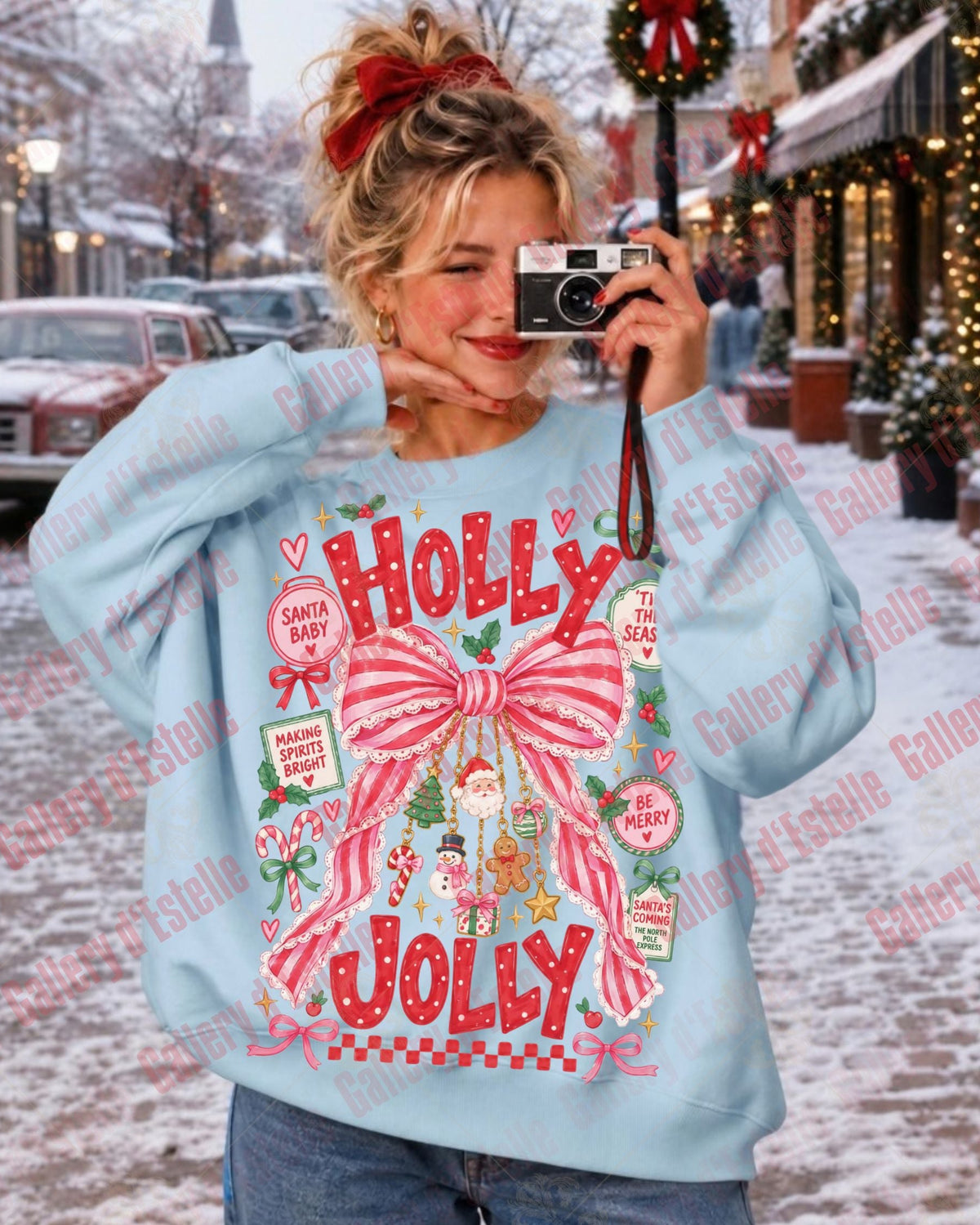 A woman takes a mirror selfie wearing a light blue crewneck sweatshirt with a large red and pink 'Holly Jolly' Christmas graphic on the front, set against a snowy holiday street scene.