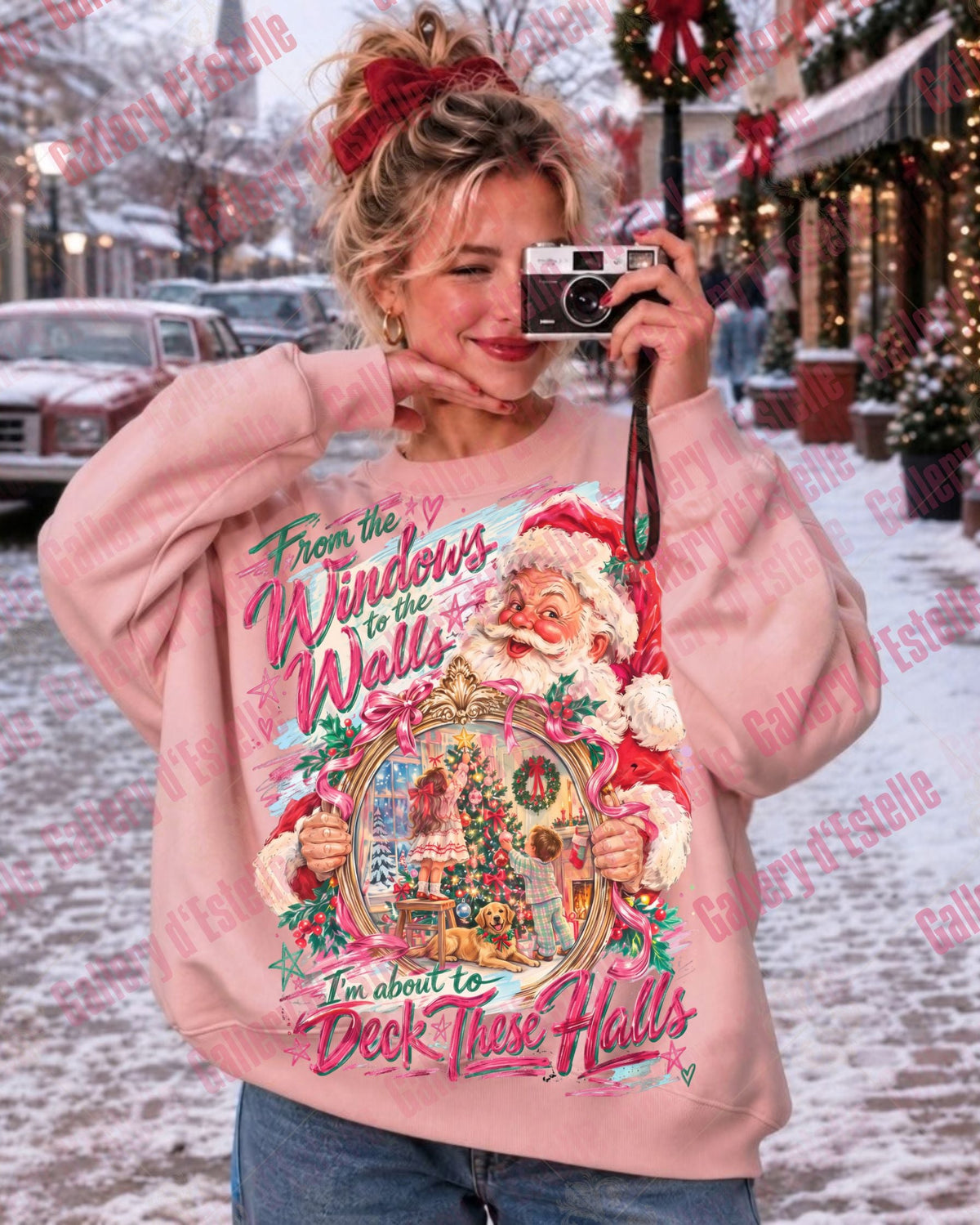 A woman wearing a light pink crewneck sweatshirt with a large, colorful Christmas graphic on the front, standing in a snowy outdoor street decorated with Christmas lights and trees.