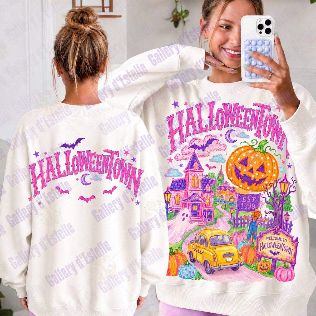 A woman shows two views of a white sweatshirt with a colorful Halloween-themed graphic that reads 'HALLOWEENTOWN.' The left view shows the back with pink lettering and bats, and the right view shows the front with a haunted house, jack-o'-lantern, moon, and car.