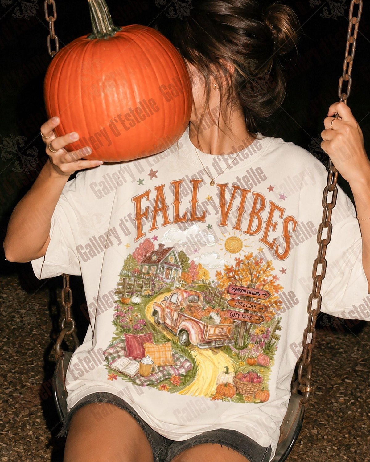 A woman sitting on a swing at night holds a large orange pumpkin to her face while wearing an oversized white graphic T-shirt that reads 'FALL VIBES' above a colorful autumn illustration. 