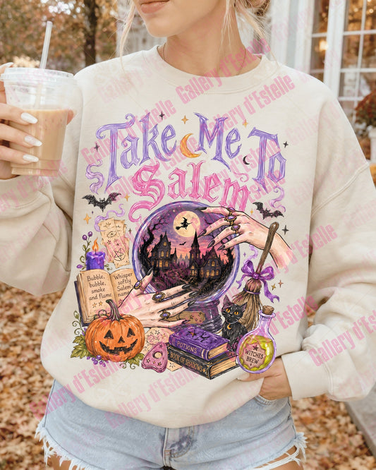 A woman wearing a cream-colored sweatshirt with a colorful 'Take Me To Salem' Halloween graphic, holding an iced coffee outdoors.