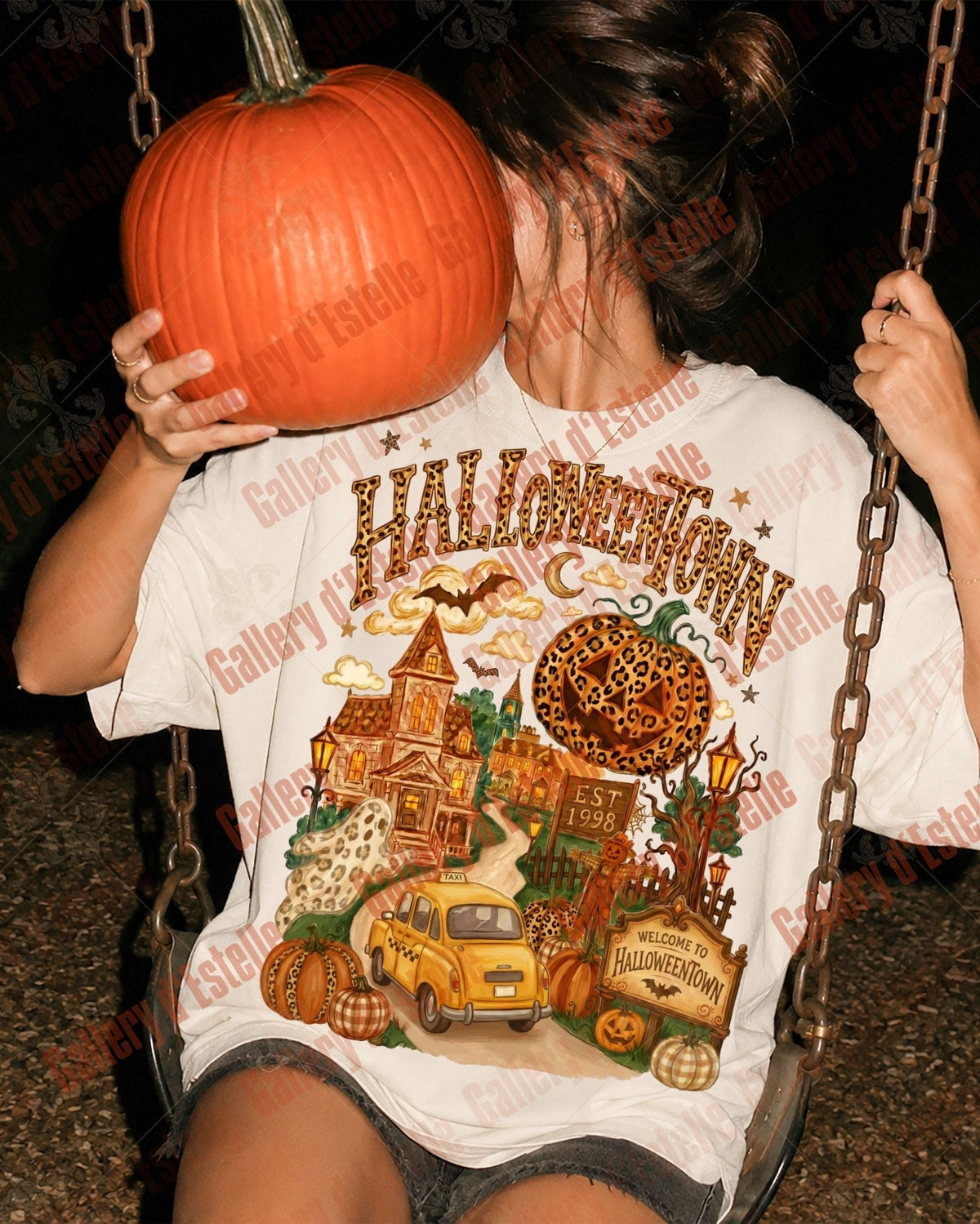 A person sitting on a swing at night wearing a white graphic t-shirt with a colorful Halloween-themed print that reads 'HALLOWEEN NIGHT'. The person is holding a large orange pumpkin in front of their face.
