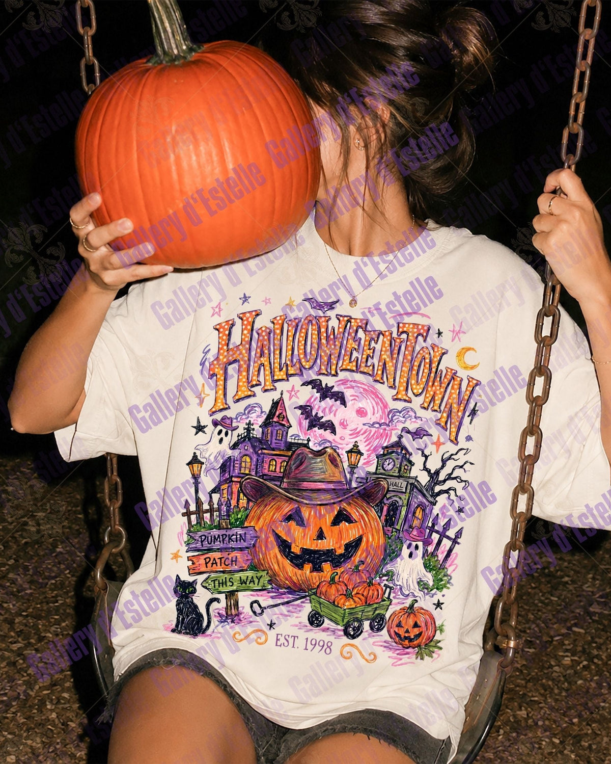 A person sitting on a swing at night wearing a white graphic t-shirt with a colorful Halloween-themed 'Halloween Town' design, holding a large orange pumpkin in front of their face.