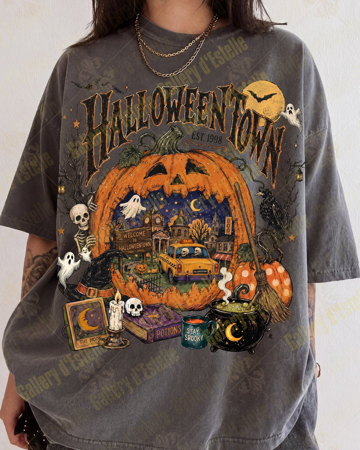 A person wearing an oversized charcoal gray graphic T-shirt with a large Halloween-themed print featuring a jack-o'-lantern, ghosts, skeletons, a cauldron, and the words 'Halloween Town'.
