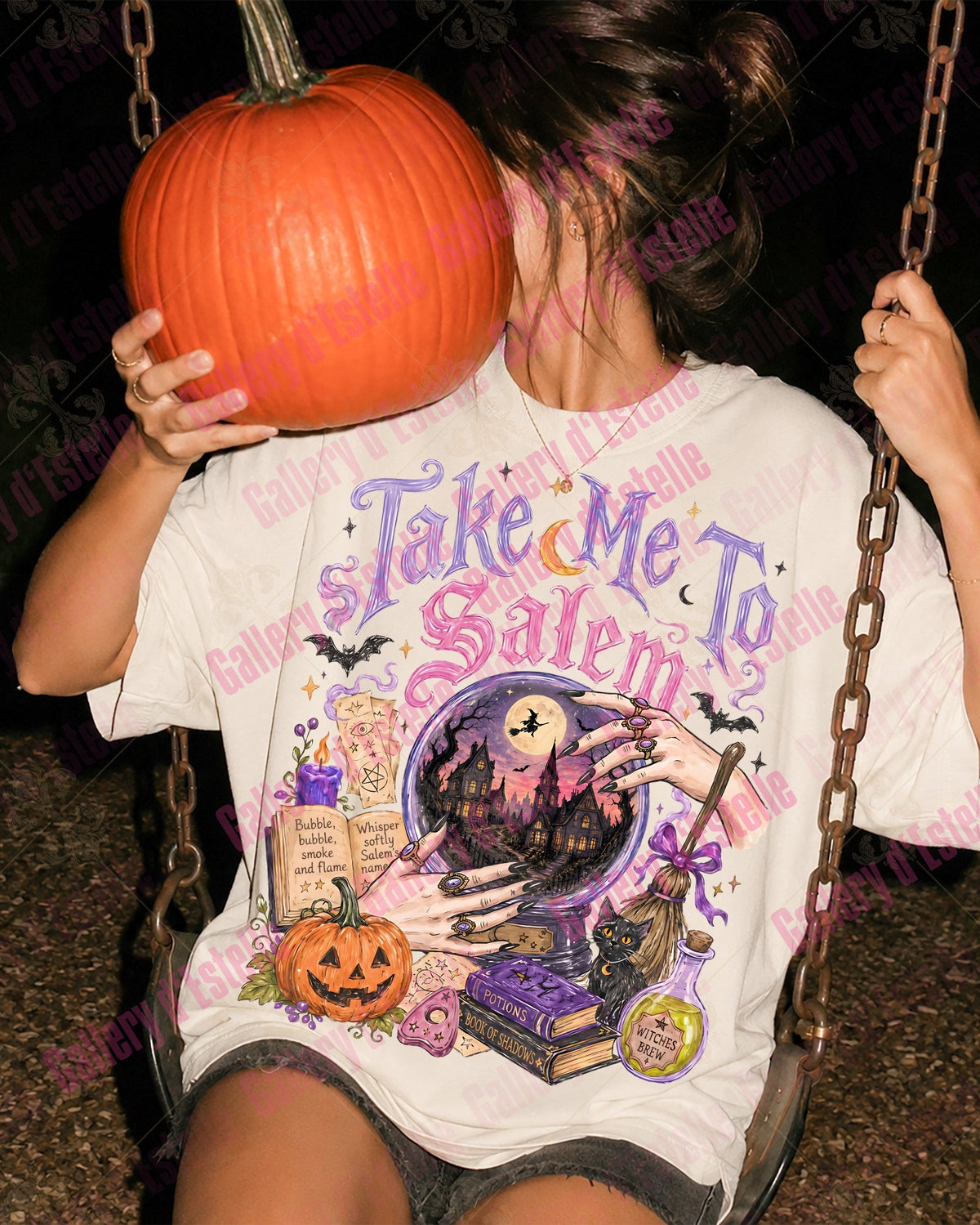 A person sitting on a swing at night wearing a white graphic T-shirt with a Halloween design that reads 'Take Me To Salem' and featuring a witch's circle, potion bottle, pumpkin, and bat motifs. The person is holding a large orange pumpkin in front of their face.