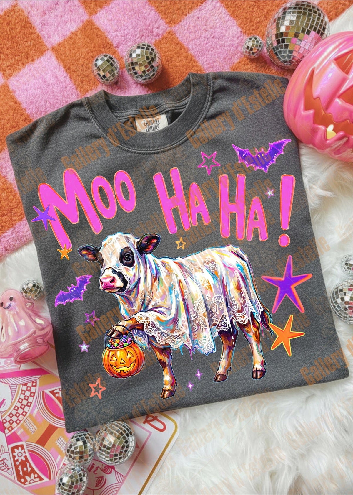 A dark gray graphic t-shirt featuring a Halloween-themed design with a cow in a poncho, a jack-o'-lantern, bats, and the text 'Moo Ha Ha Ha!' in pink. The shirt is displayed in a flat-lay arrangement with various seasonal props including a jack-o'-lantern, mini mirrors, and a pink and orange checkered blanket.