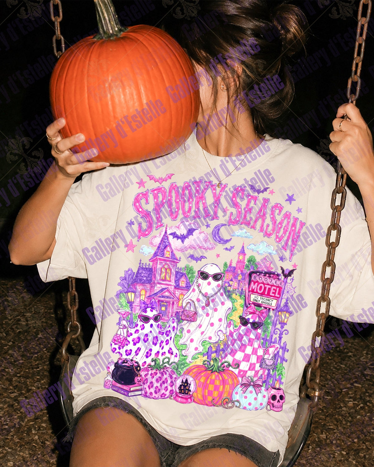A person sitting on a swing at night holds a large orange pumpkin in front of their face while wearing a white graphic T-shirt with a colorful Halloween design that reads 'SPOOKY SEASON' and includes a haunted house, glam ghosts and pumpkins.