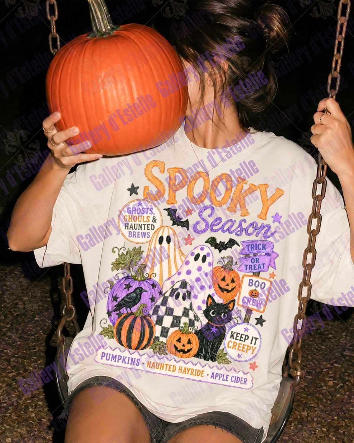 A person sitting on a swing at night wearing a white graphic T-shirt with Halloween-themed artwork and the words 'Spooky Season' in orange and purple. The person is holding a large orange pumpkin in front of their face.