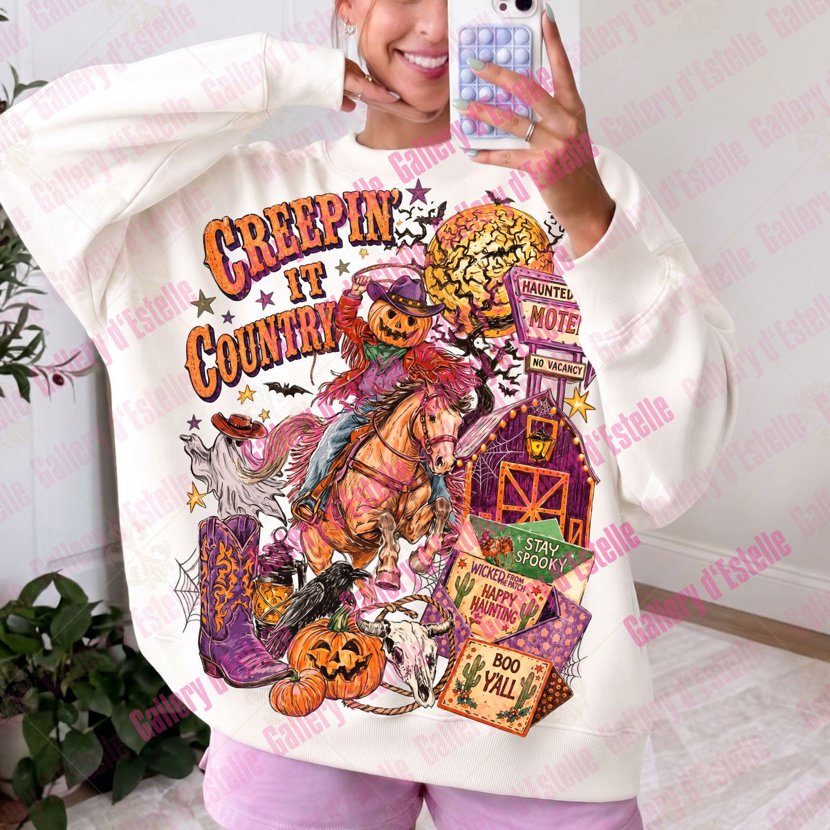 A woman takes a mirror selfie wearing a cream-colored sweatshirt with a large, colorful Halloween-themed graphic that reads 'CREEPIT COUNTRY' and features pumpkins, a witch, a haunted barn, and a cactus.