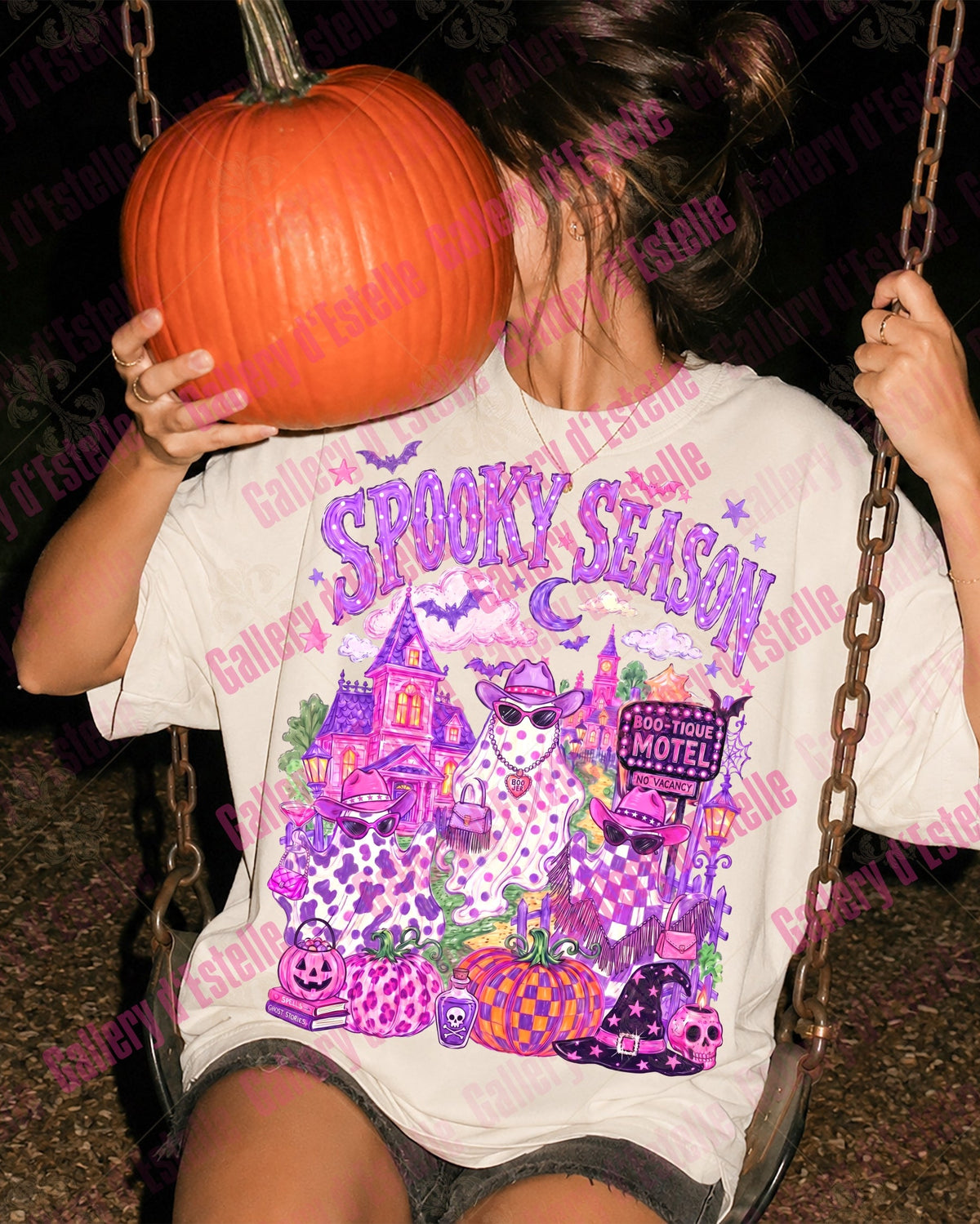 A person sitting on a swing at night wearing a white graphic t-shirt with a purple and pink 'Spooky Season' Halloween design. The person is holding a large orange pumpkin in front of their face.