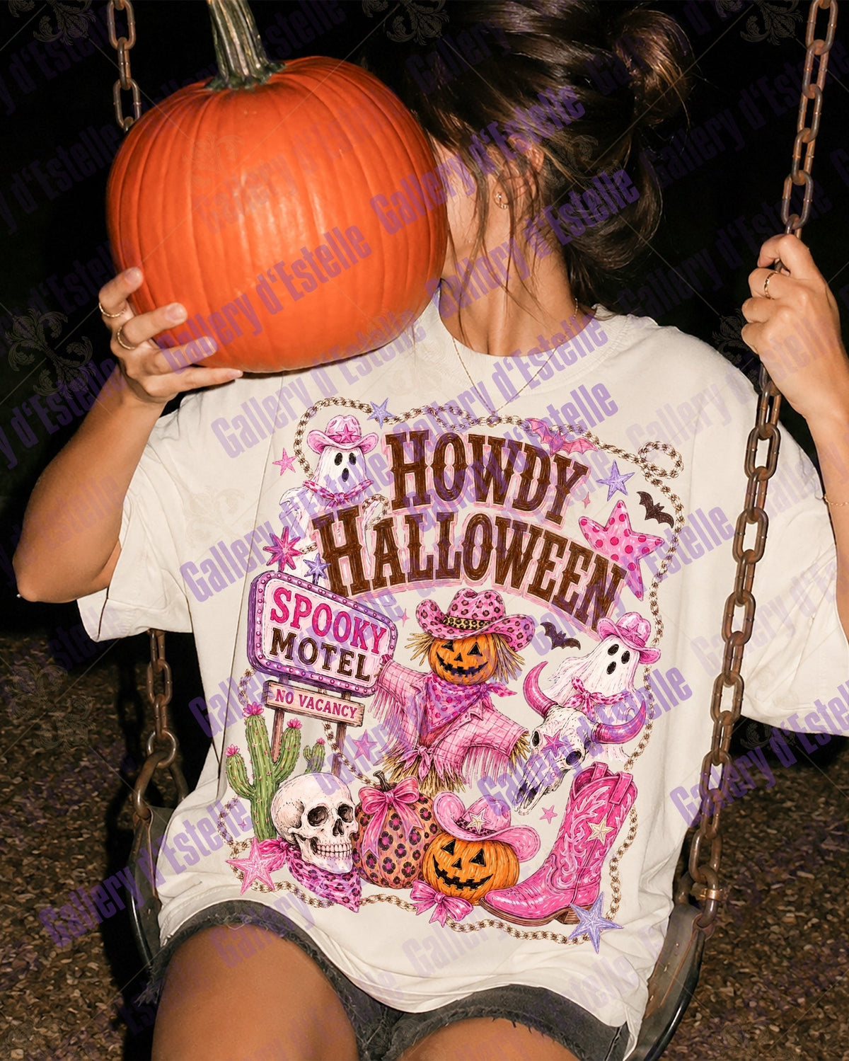 A person sitting on a swing at night wearing a white graphic T-shirt with a colorful Halloween-themed print that reads 'HOWDY HALLOWEEN' and 'SPOOKY MOTEL'. The person is holding a large orange pumpkin over their face.