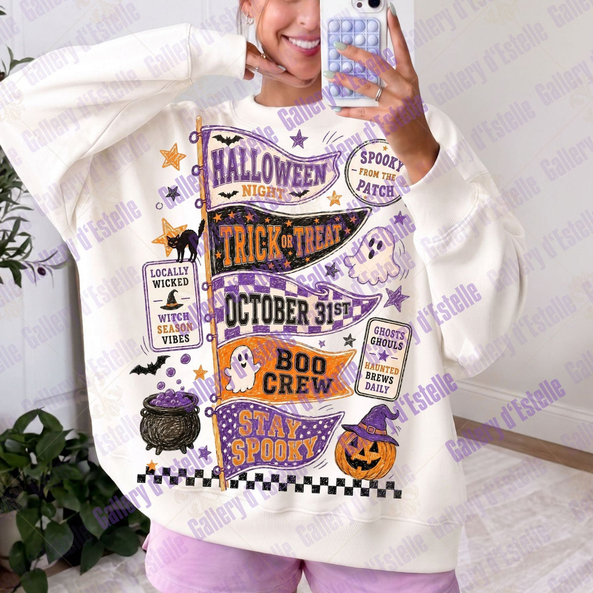 A person taking a mirror selfie wearing an oversized white sweatshirt with a large purple, orange, and black Halloween-themed graphic print featuring text like 'Halloween Night', 'Trick or Treat', 'Stay Spooky', and 'Boo Crew'.