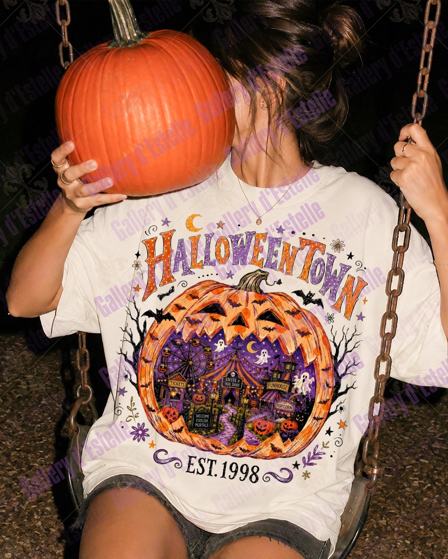 A person sitting on a swing at night wearing a white graphic t-shirt with a Halloween-themed 'Halloween Town' design, holding a large orange pumpkin in front of their face.