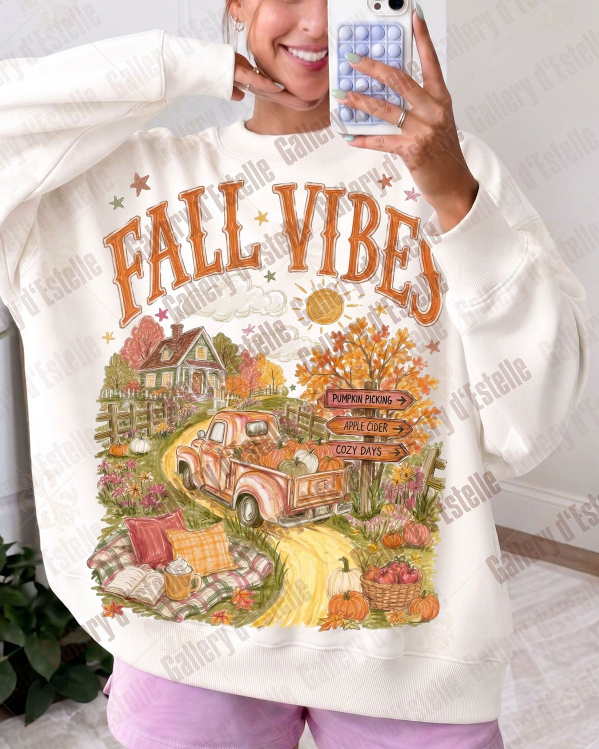 A woman taking a mirror selfie wearing a cream-colored sweatshirt with a large 'FALL VIBES' graphic print featuring an autumn truck, house, and pumpkins.